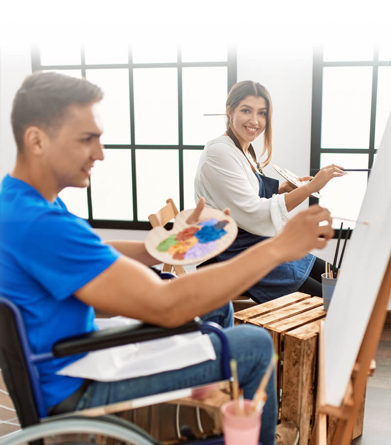 Ember Support Services - Community Access & Social Inclusion page, Community Access NDIS hero image of a young woman and a man in a wheelchair painting a picture each in a white studio.